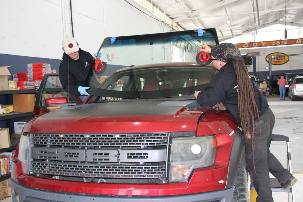 Two technicians installing a windshield on a red Ford truck in an auto shop.