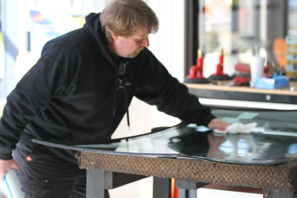 Man installing a windshield in an auto repair shop, using a cloth and tools on a workbench.