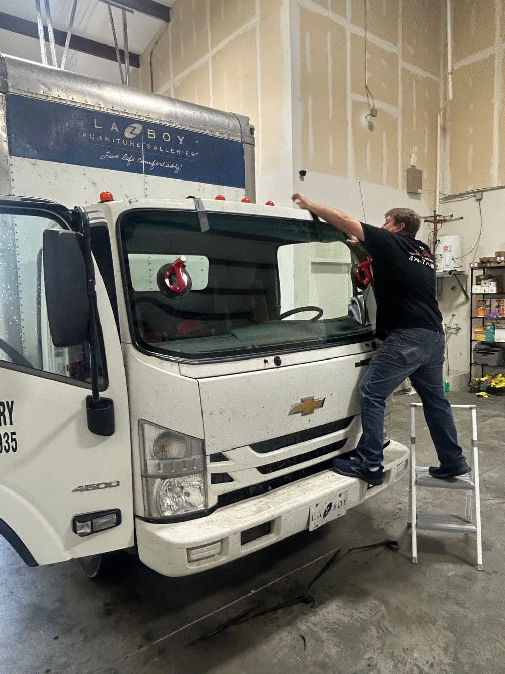 Man cleaning the windshield of a Chevrolet delivery truck using a ladder inside a warehouse.