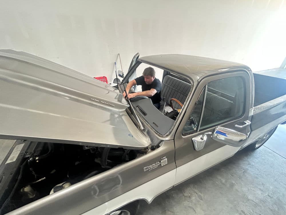 Mechanic working on a classic gray pickup truck in a garage with the hood open.